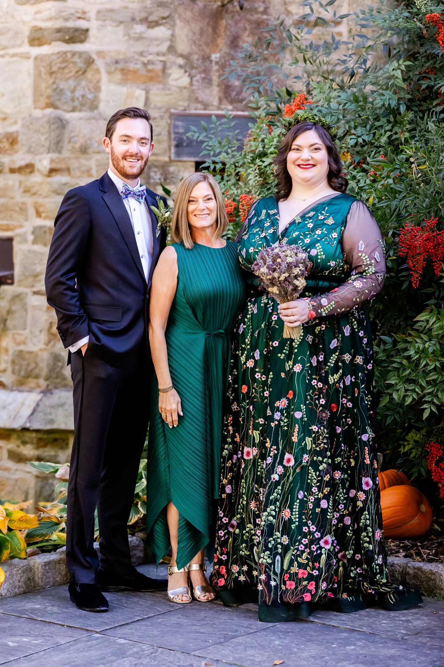 Three wedding attendees pose together against a stone wall with autumn decorations.