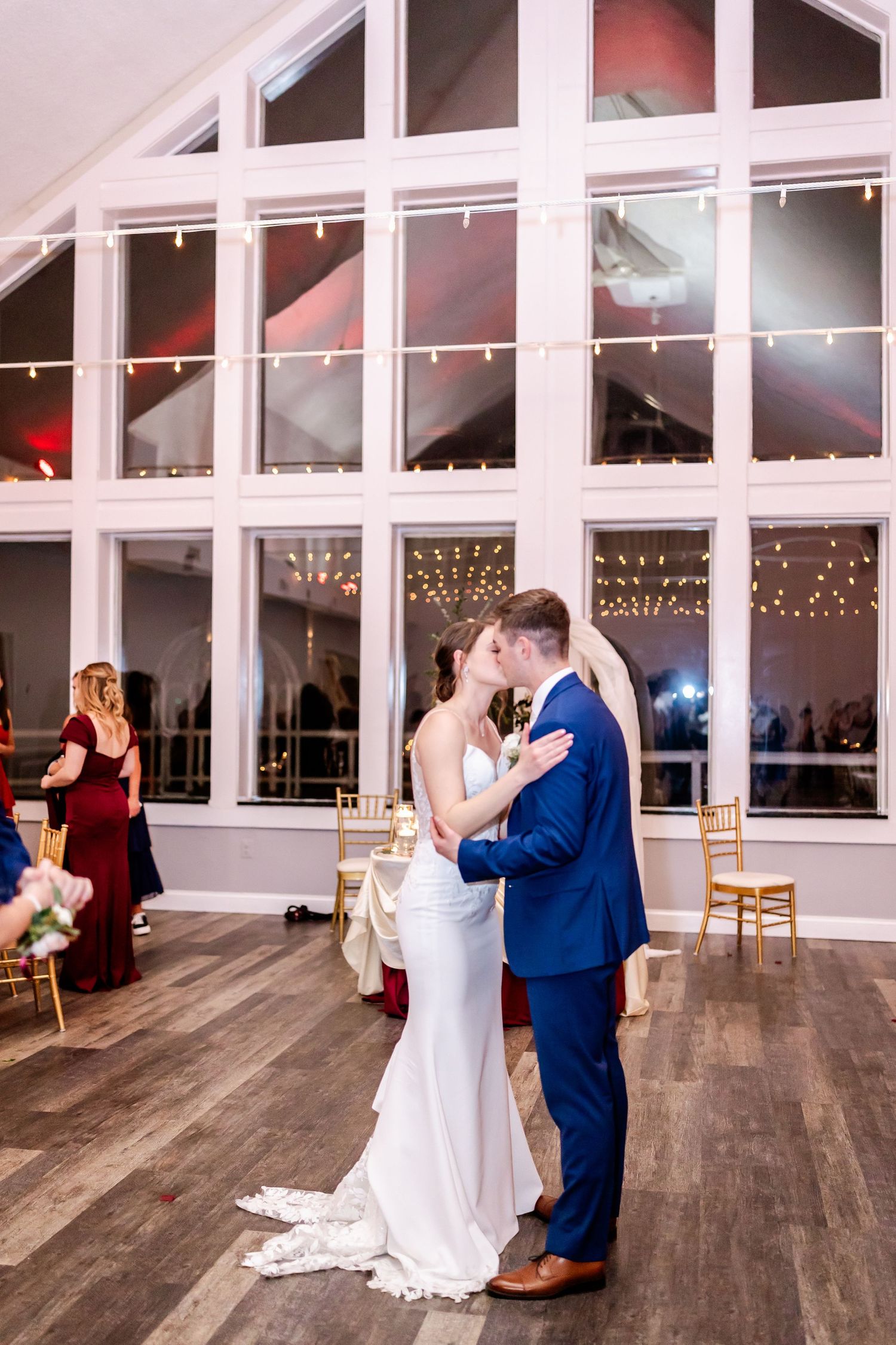 Couple sharing their first dance in an elegant white venue with large windows and string lights.