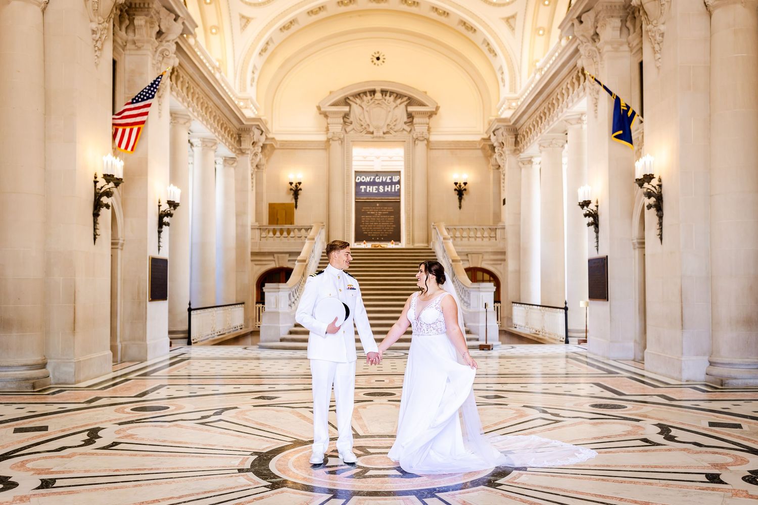 Couple stands in an ornate marble hall with American flag and classical architecture details.