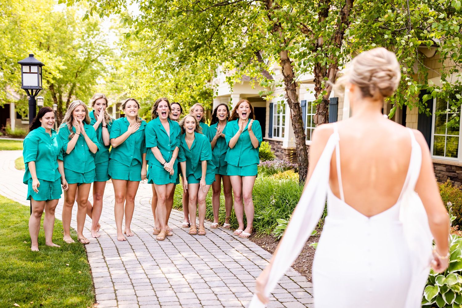 Bridesmaids in matching teal robes react with excitement as they see bride in white dress on garden pathway.