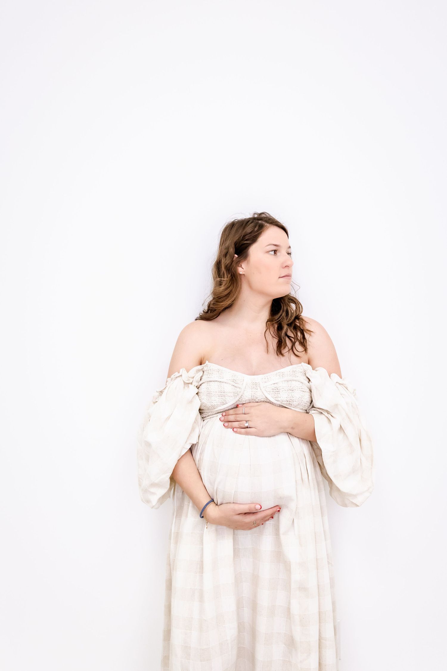 A maternity portrait of someone in a flowing white off-shoulder dress against white background.