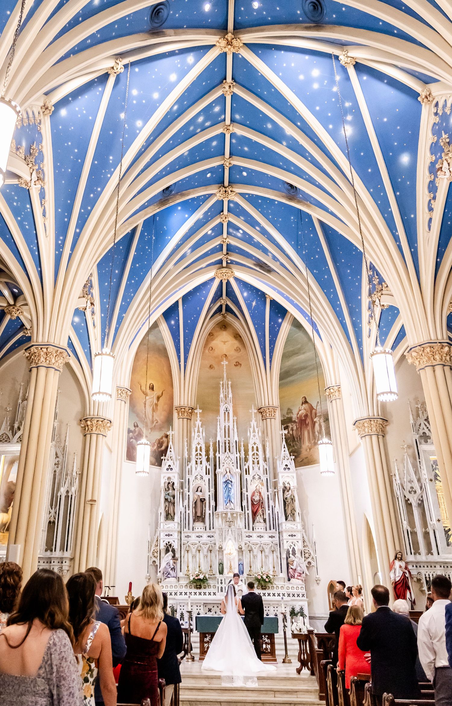 Stunning cathedral interior with Gothic vaulted blue ceiling and ornate white architectural details during ceremony.