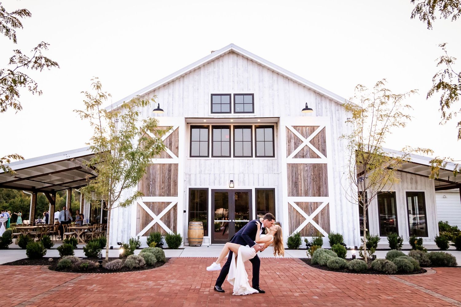 A modern white barn wedding venue with rustic crossed-beam details and brick patio surrounded by lush landscaping.