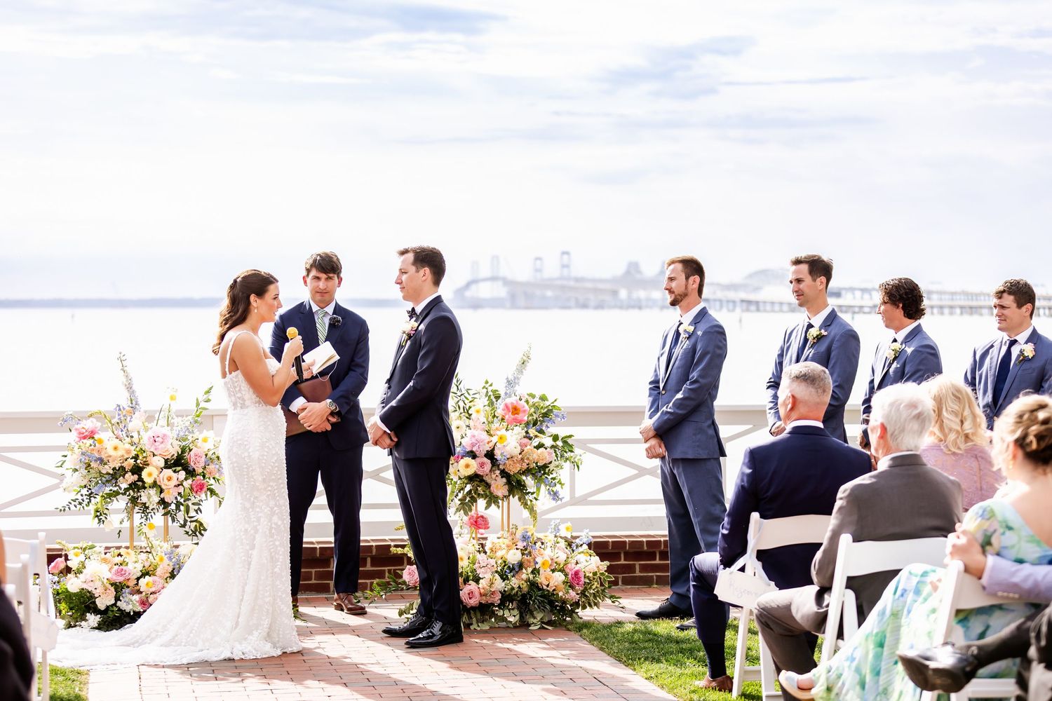 Outdoor waterfront wedding ceremony with floral arrangements and guests seated on white chairs.