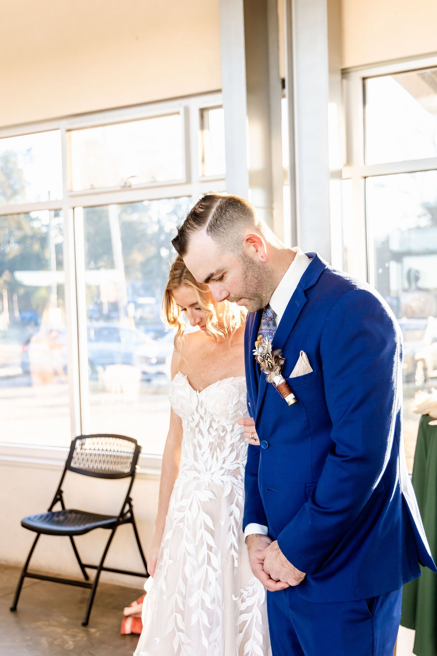 A tender wedding moment between a couple, with the bride in white lace and groom in royal blue suit near sunlit windows.