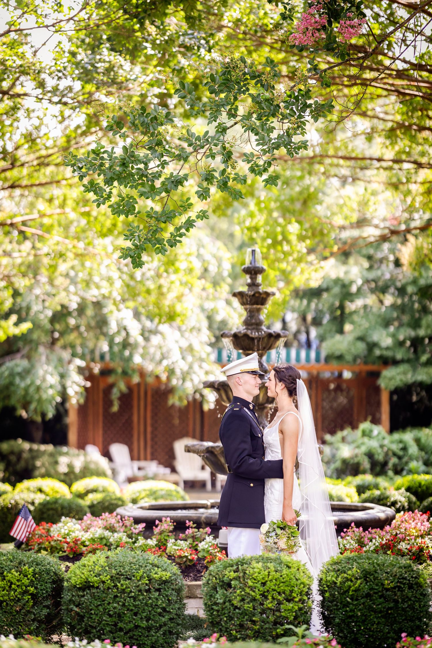 A couple shares an intimate moment by an ornate garden fountain surrounded by lush greenery and flowers.