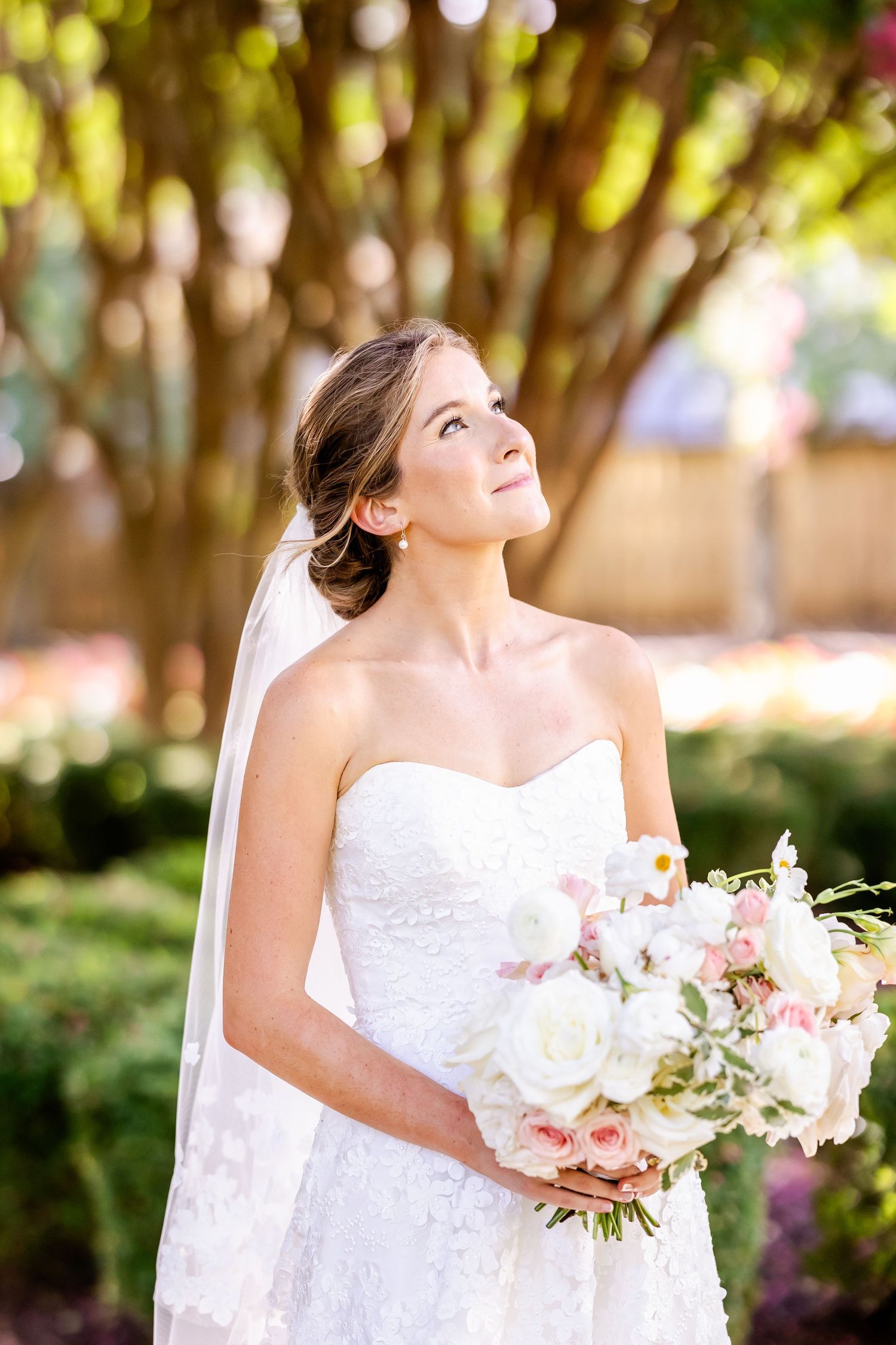 A bride in a white wedding dress holds a pink and white bouquet in an outdoor garden setting.