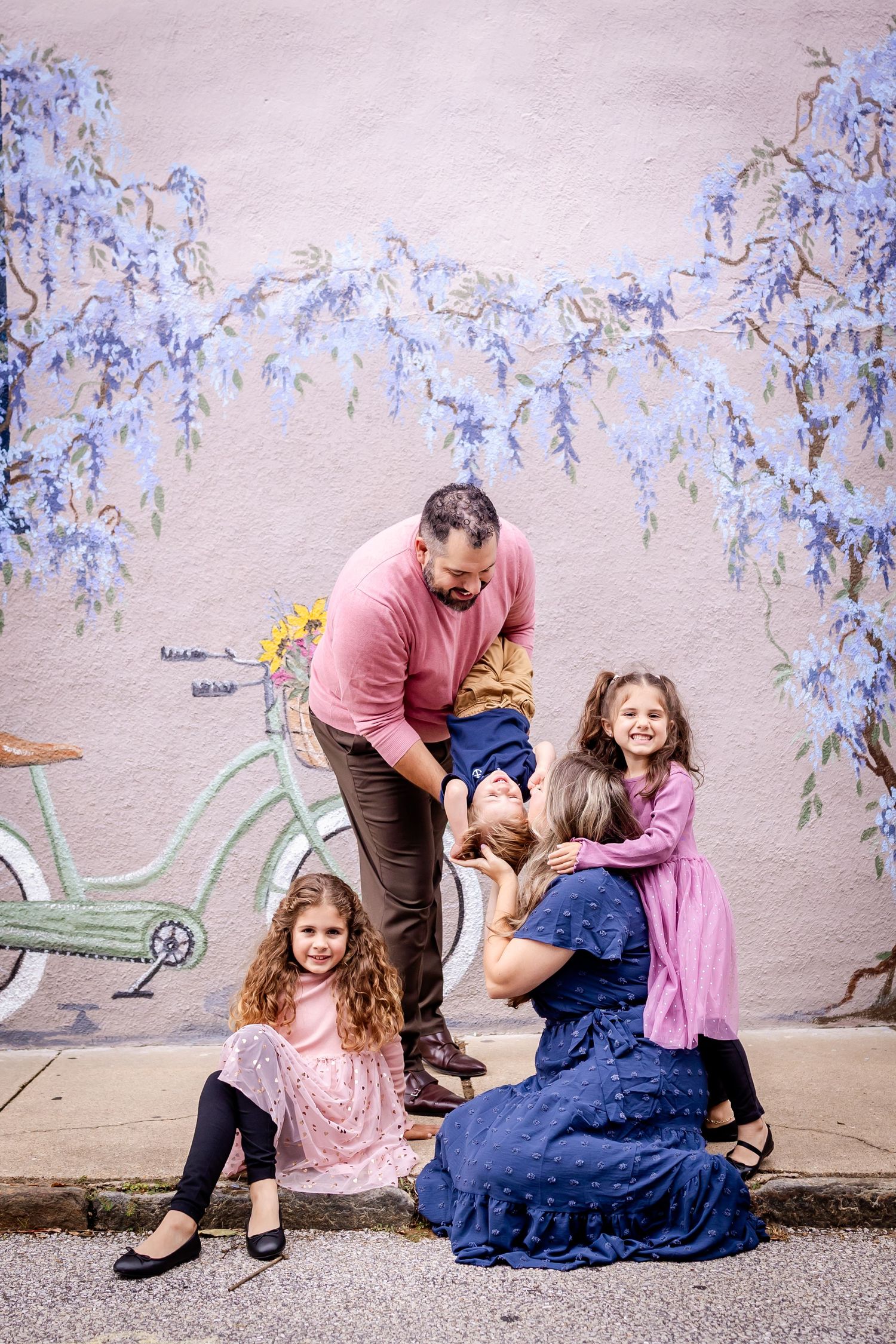 A family plays and interacts together in a playful photo sequence against a wall with hanging blue flowers.