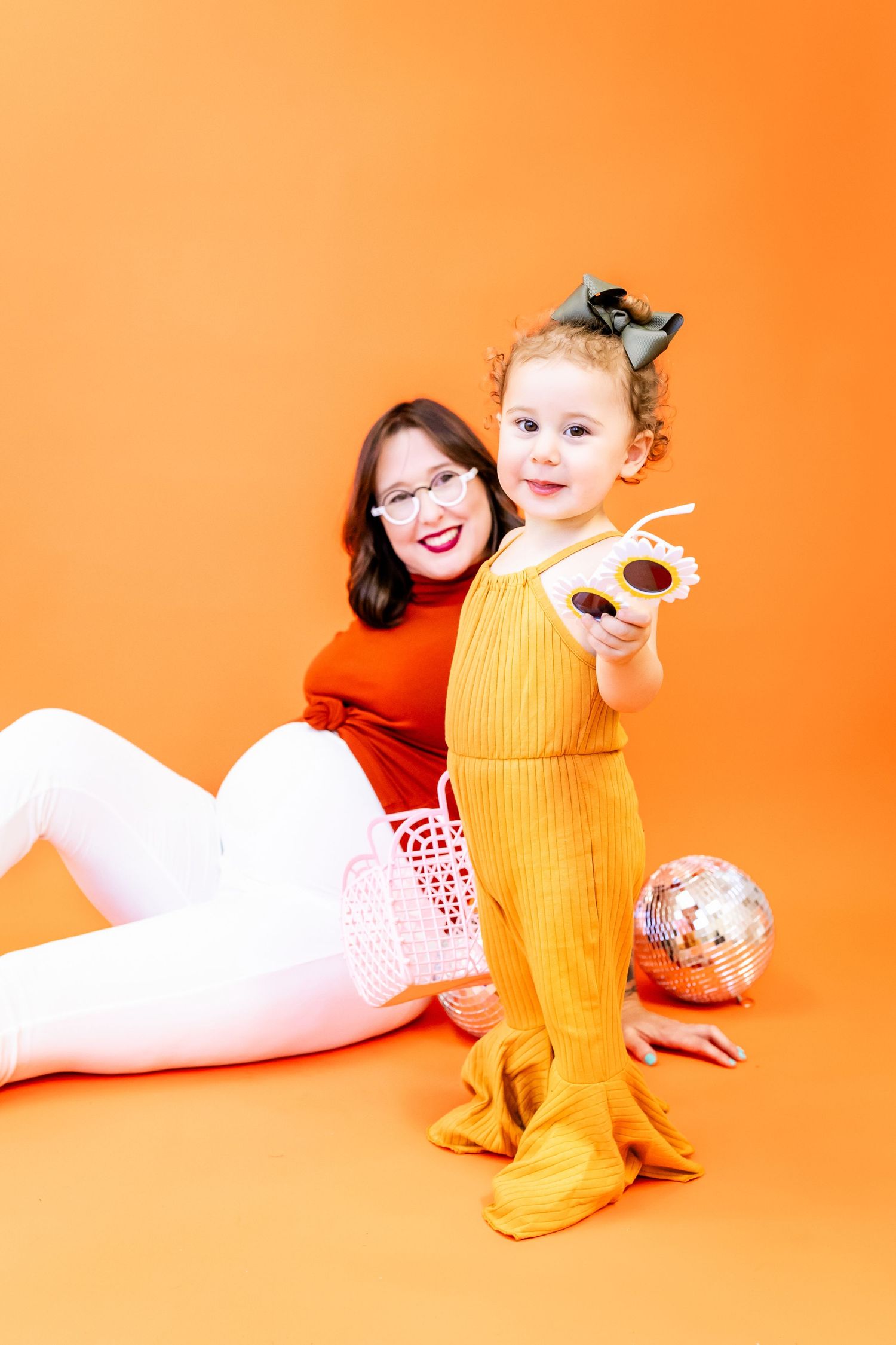 A baby in a yellow knit romper plays on a bright orange backdrop while an adult in red reclines nearby.