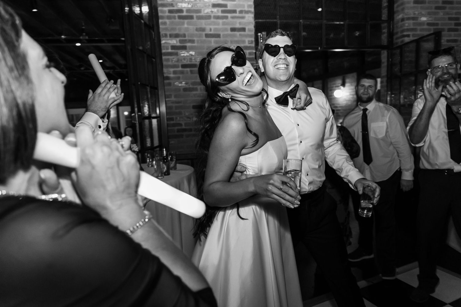 Wedding guests in sunglasses laugh and dance at a lively reception, with one woman leaning back in a white dress as friends cheer nearby.