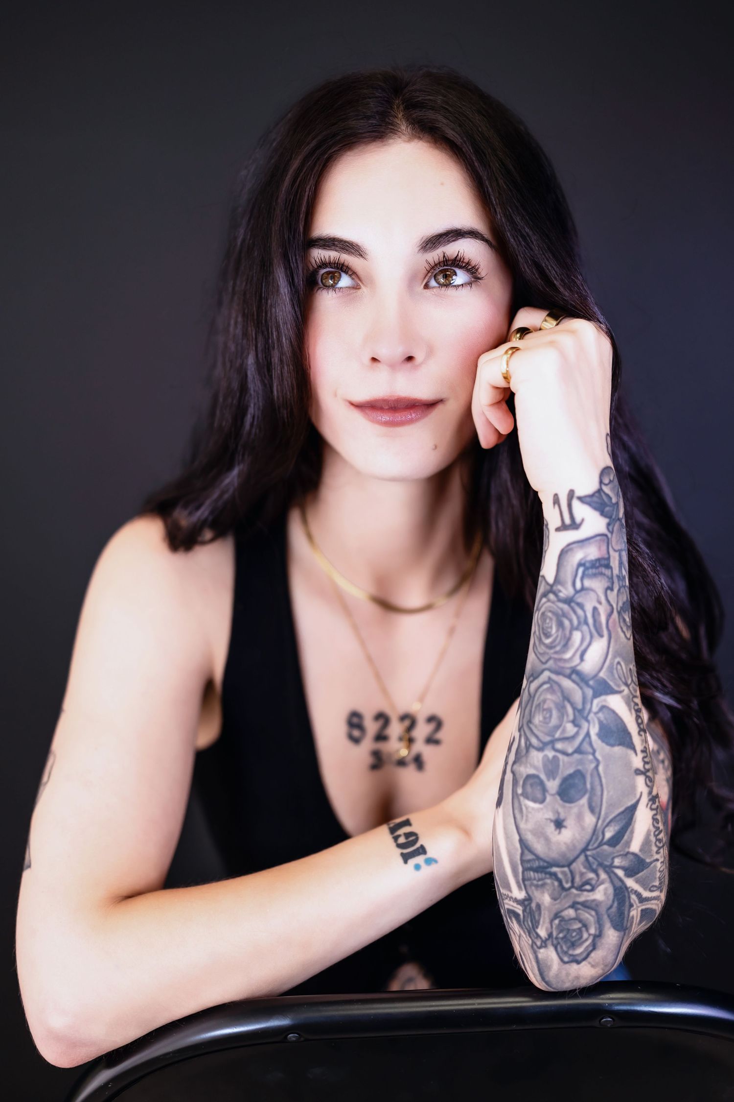 A tattooed woman with long dark hair poses thoughtfully, resting her cheek on her fist against a dark background.