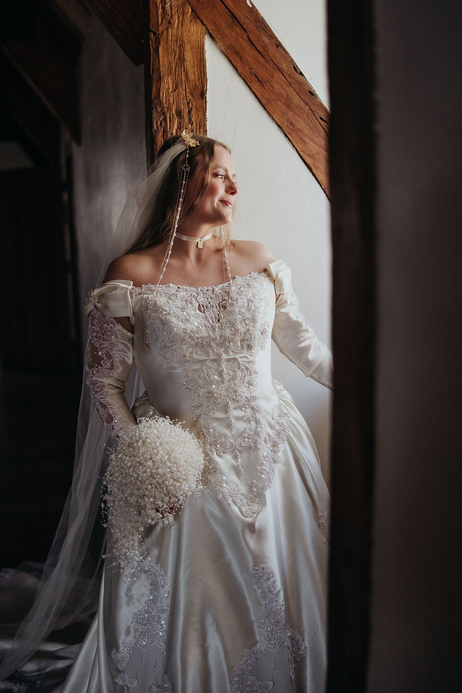 Bride in elegant off-shoulder lace wedding gown with veil and bouquet, standing by a window in a rustic venue.