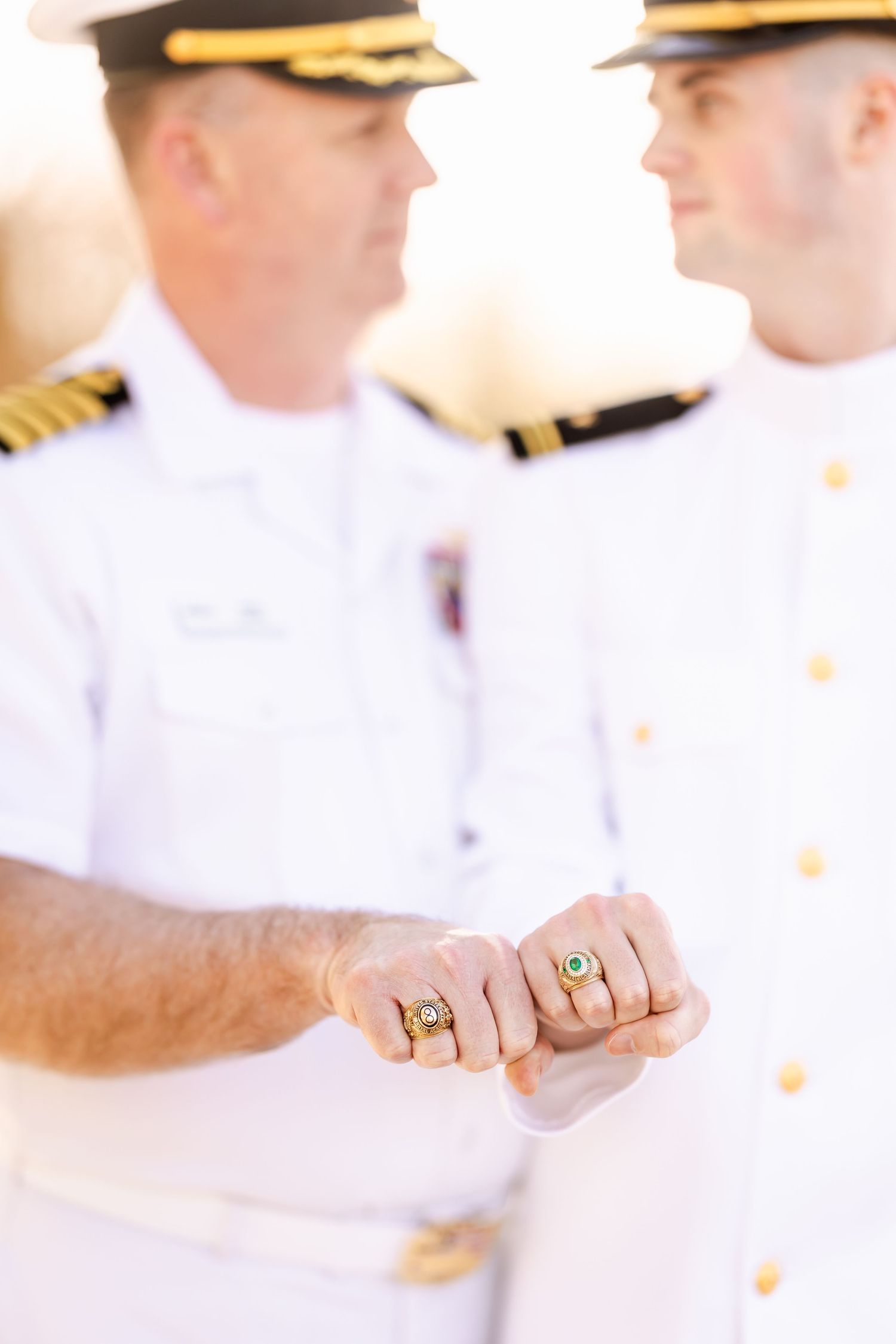 Two naval officers in white uniforms display gold class rings, fist bumping to show off their matching insignia rings.