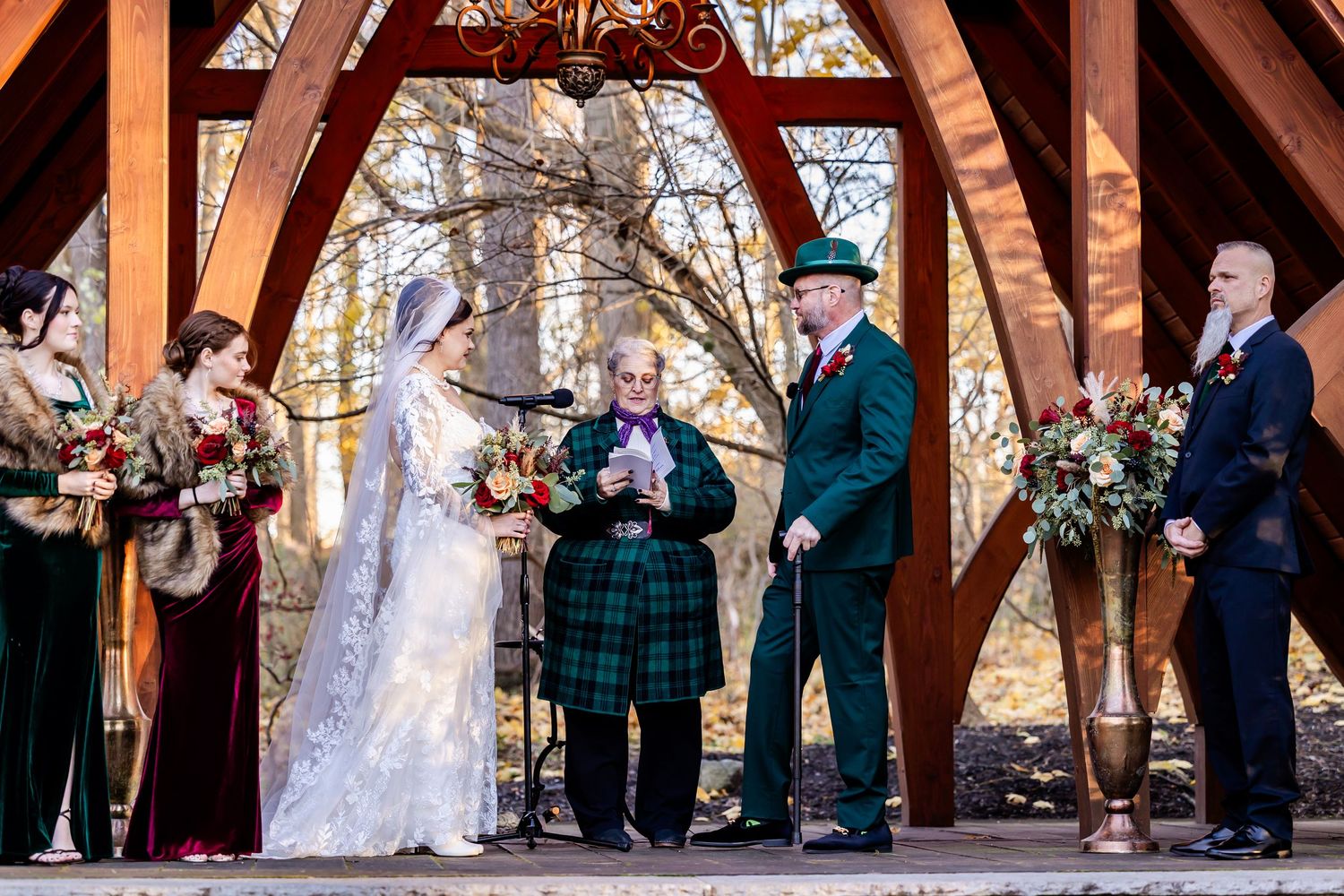 A wedding ceremony takes place under a wooden pavilion with attendants and officiant.