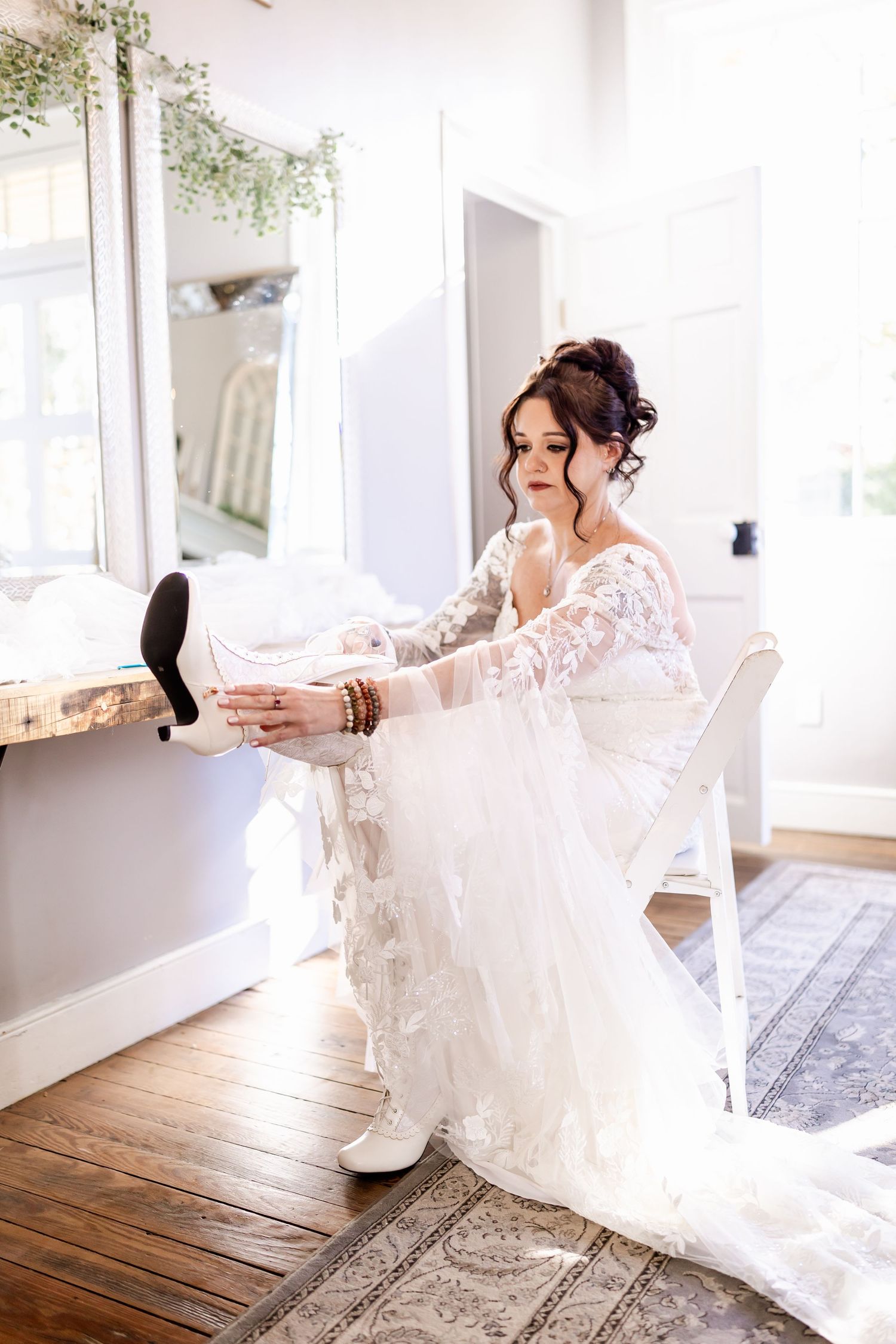 A stunning lace wedding dress being worn in a bright, sunlit dressing room with hardwood floors.