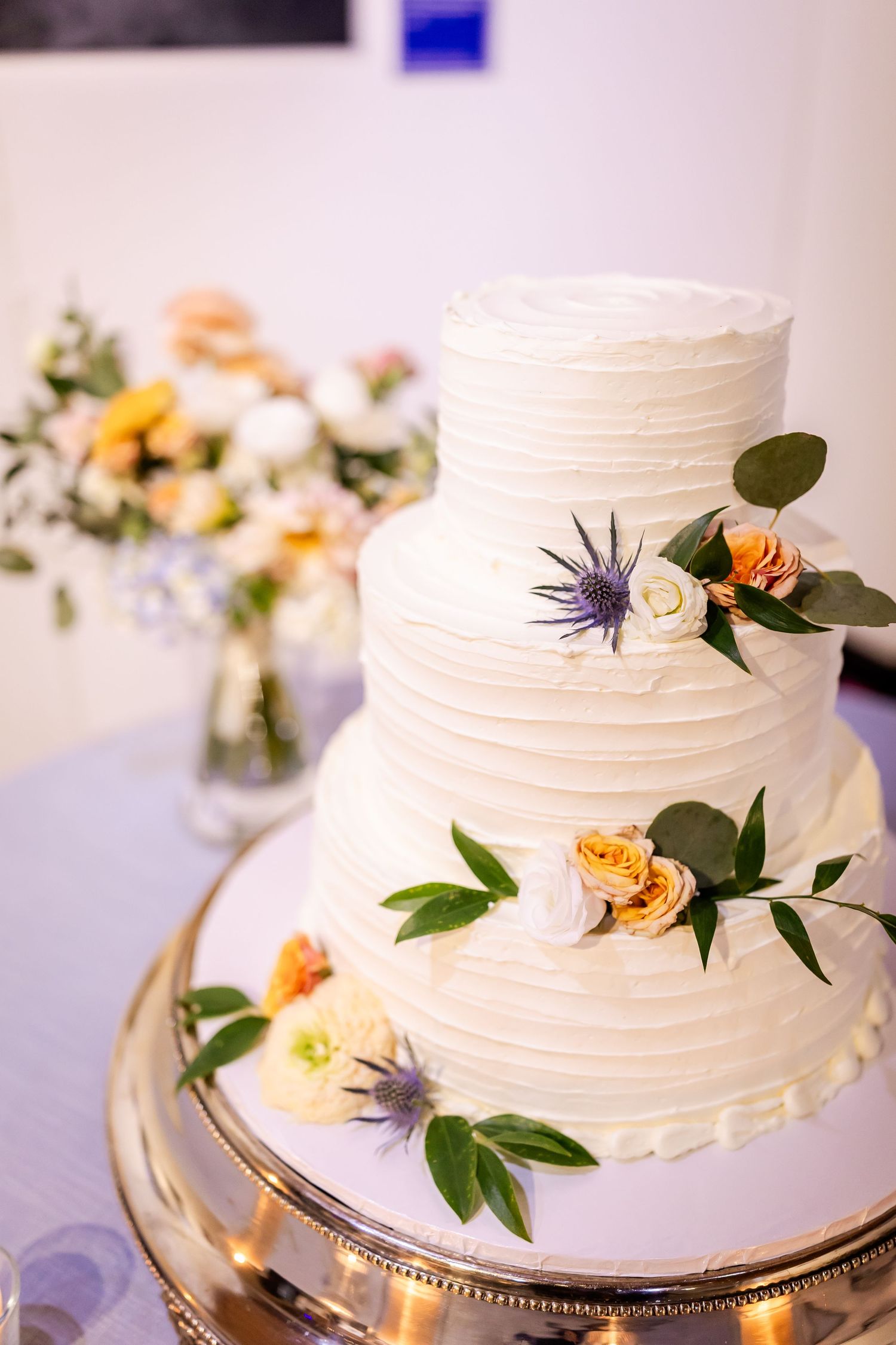 Three-tier white wedding cake with roses, greenery, and blue thistle on a silver stand.