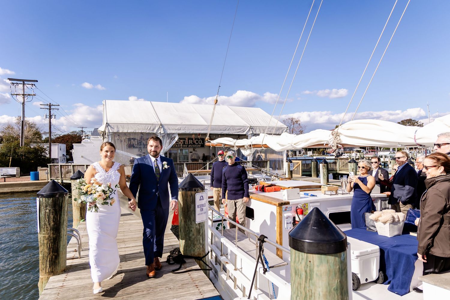 Bride and groom walk hand-in-hand along a sunny marina dock, guests applauding nearby, with sailboats and a tent venue behind.