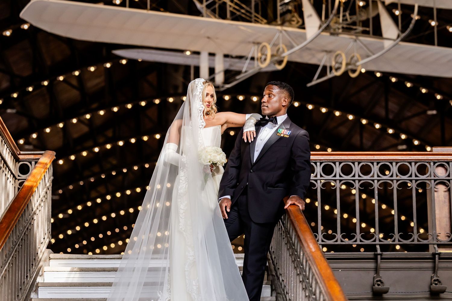 Bride and groom pose elegantly on a staircase inside a vintage venue with string lights and a historic airplane display.