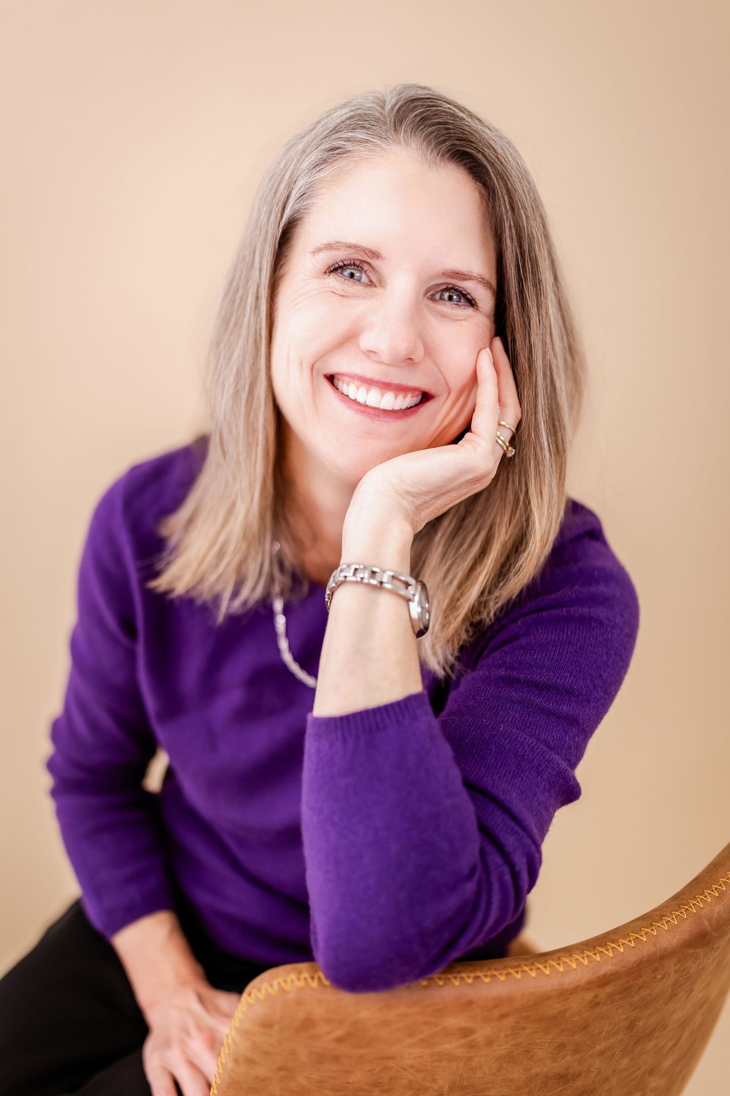 Smiling middle-aged woman with gray hair in purple sweater, resting chin on hand, seated in brown chair.