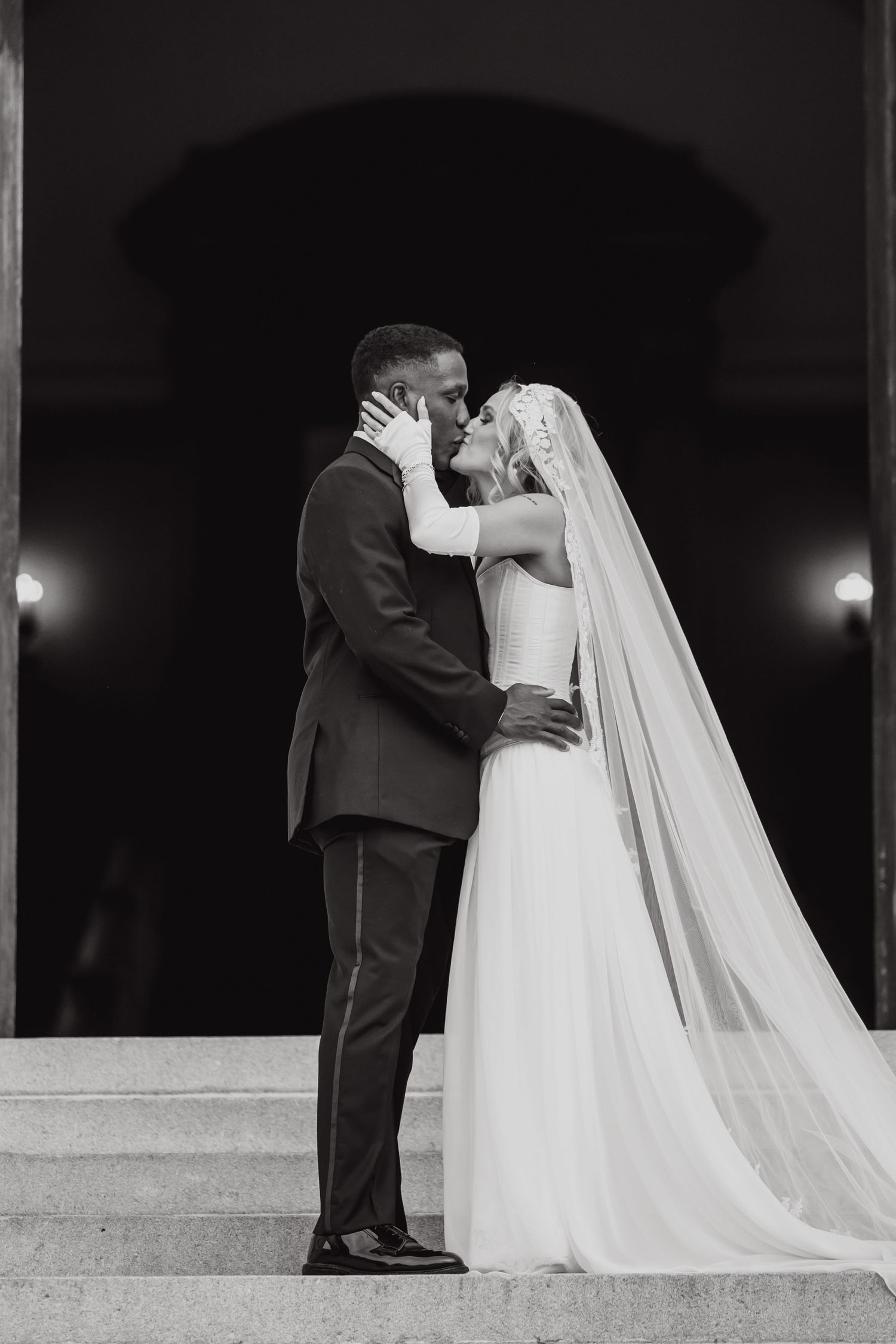 Black and white photo of a bride and groom sharing a kiss on stone steps, with the bride's veil flowing elegantly.