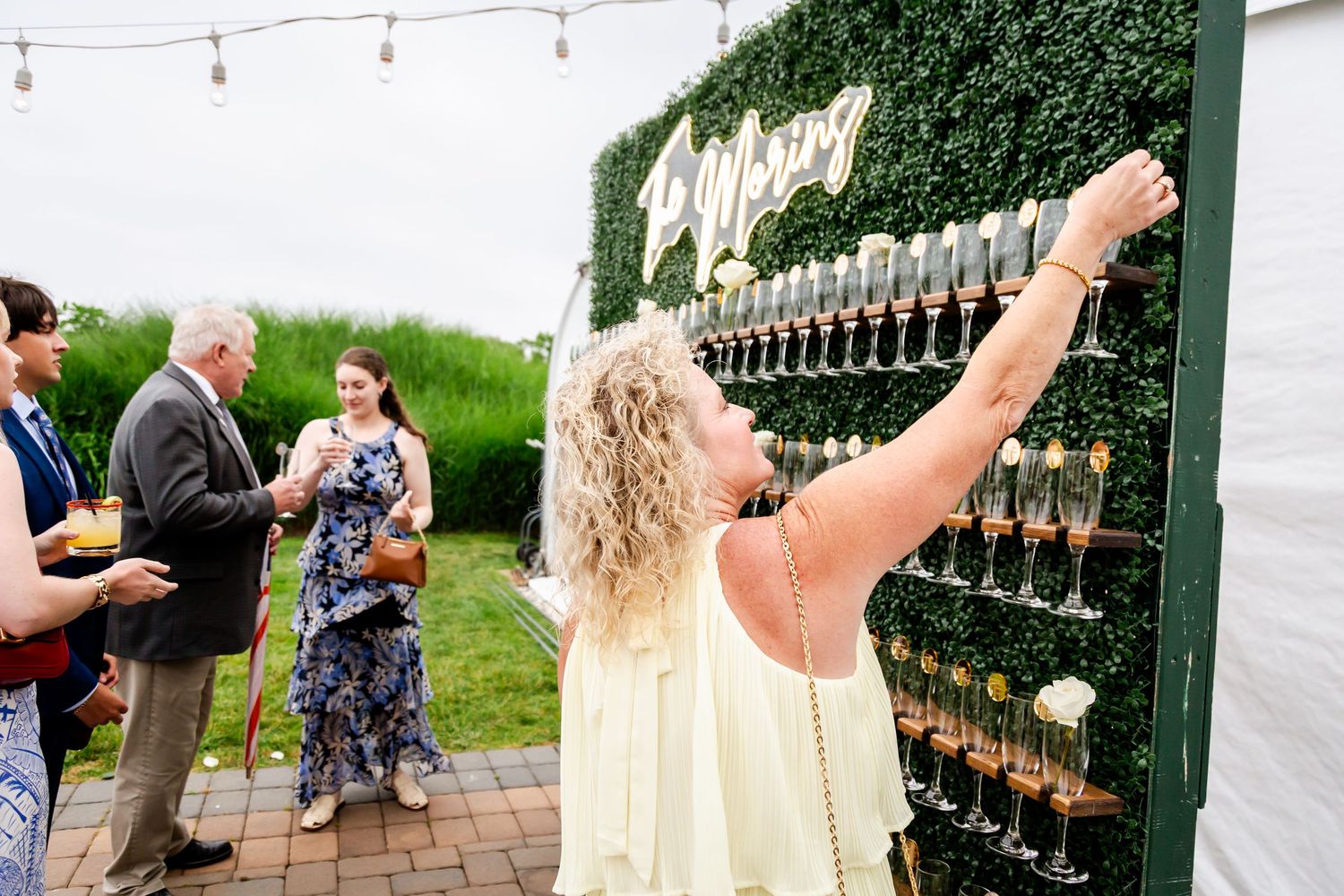 Wedding reception setup with string lights and champagne glasses against a green hedge wall.