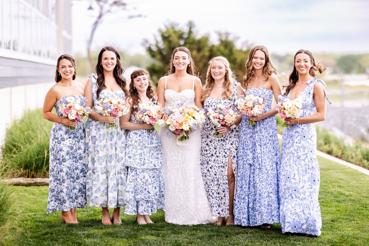 Bridesmaids in blue floral dresses hold bouquets while posing outdoors with bride.