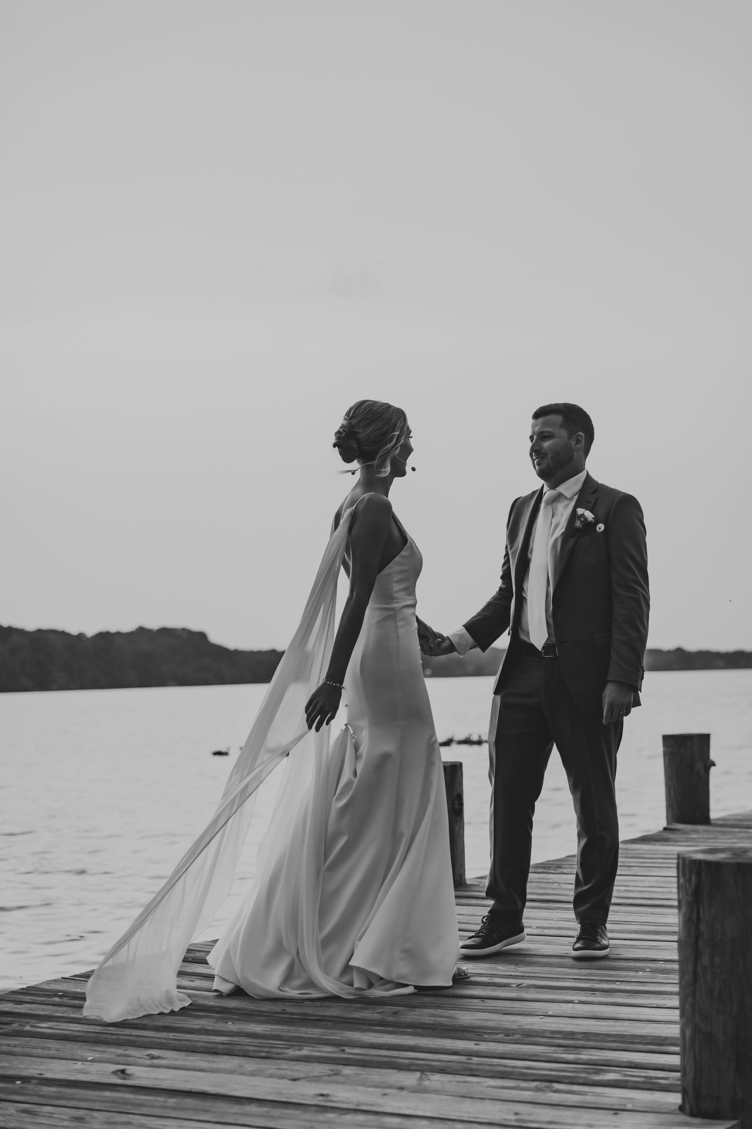 A black and white wedding photo of a couple standing on a wooden dock by a serene lake at sunset.