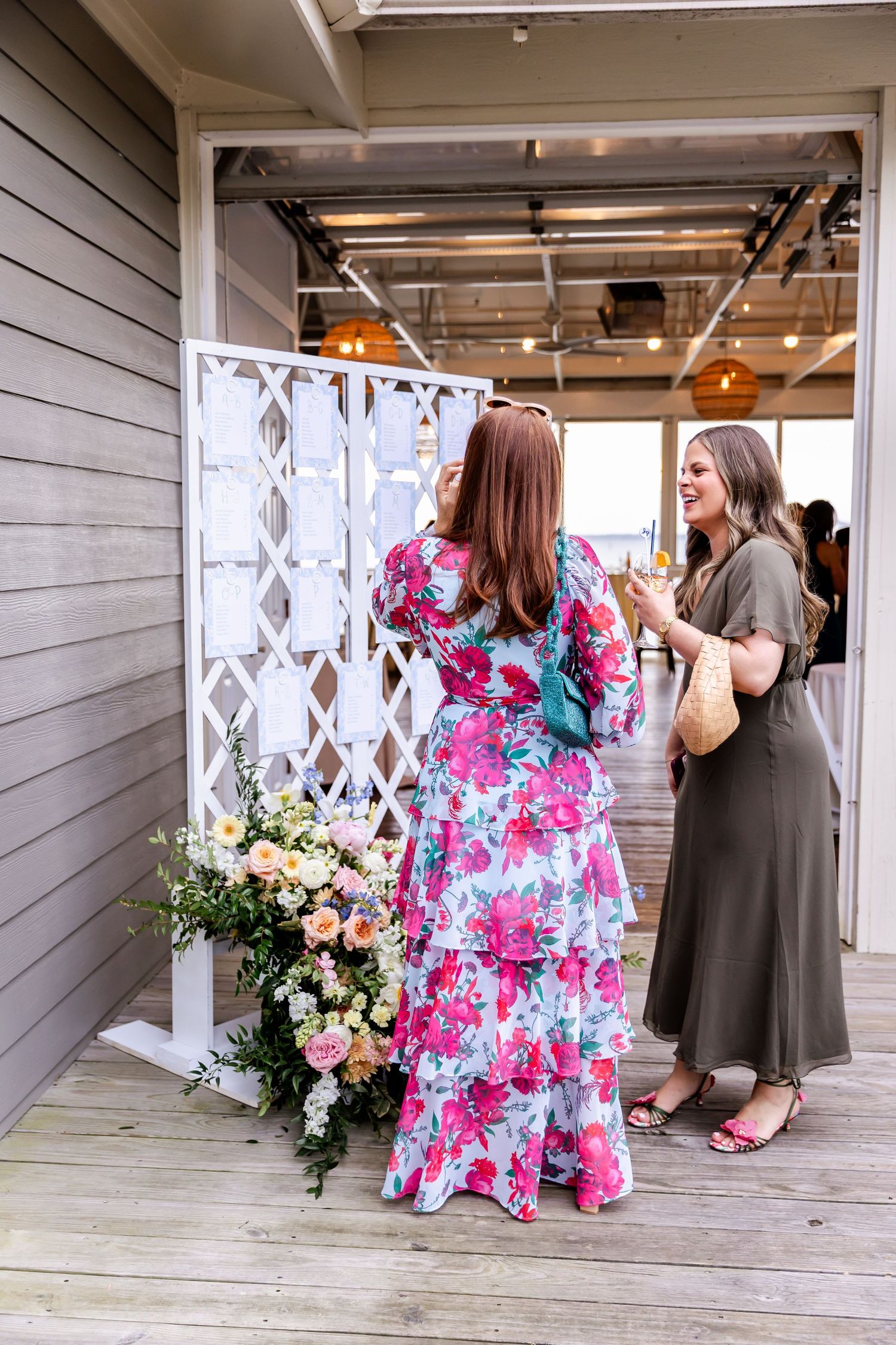Bridesmaids in bright floral dresses sit and chat on a white porch during wedding celebrations.