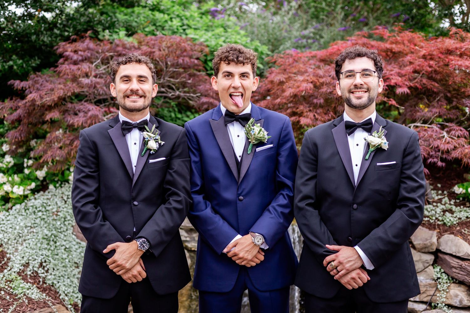 Three groomsmen in tuxedos pose in front of Japanese maple trees at a garden wedding.