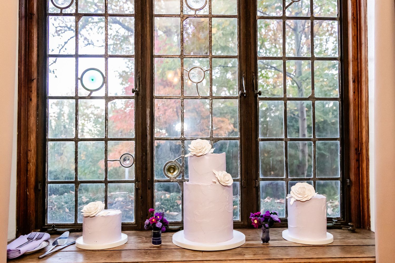 Three elegant white wedding cakes displayed on a windowsill with ornate leaded glass windows.