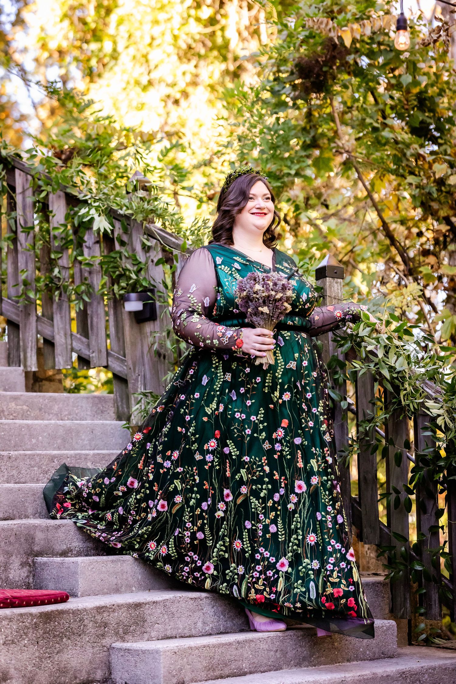 A person in a dark green floral dress with lace sleeves stands on stone steps surrounded by autumn foliage.