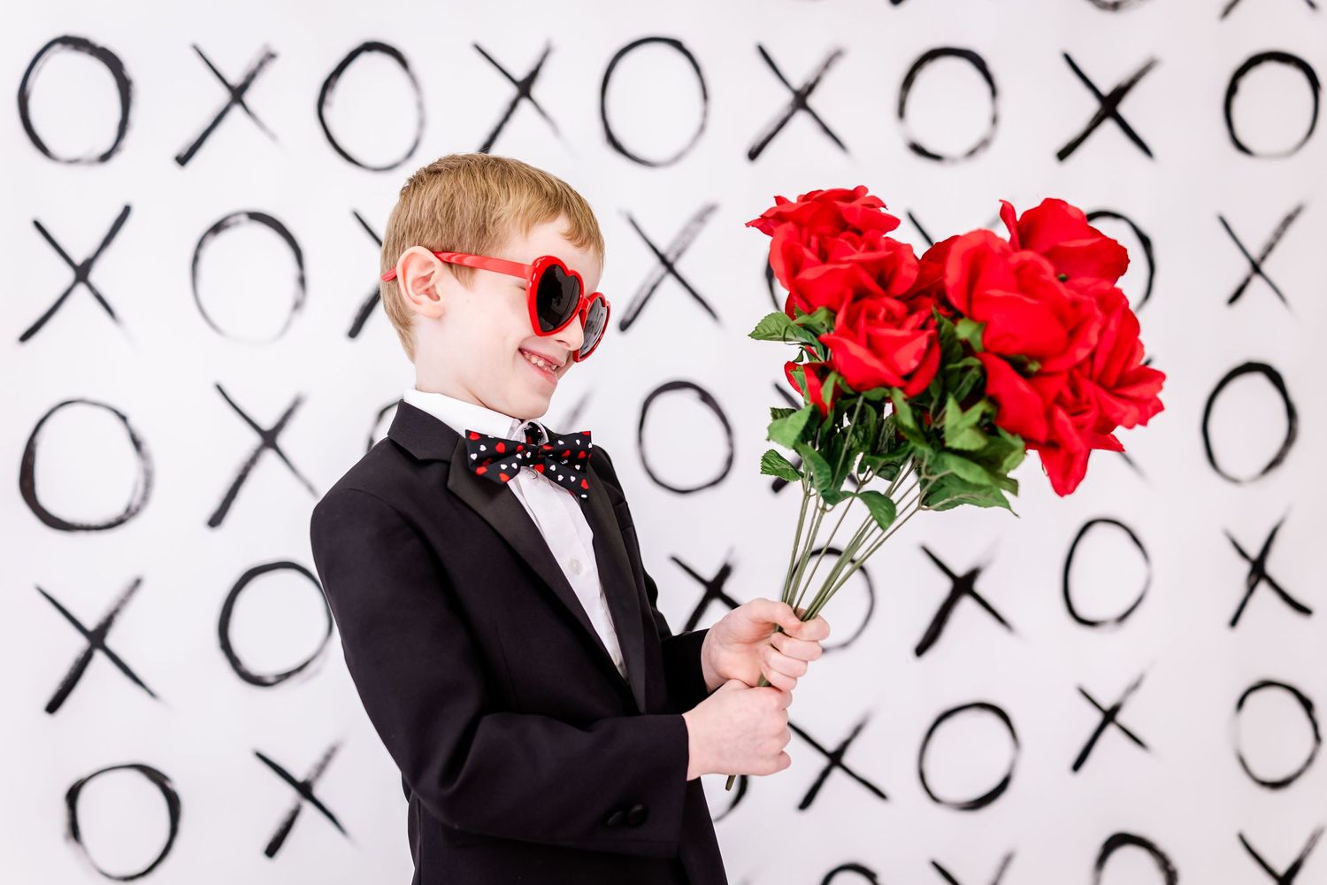 Someone in a black tuxedo and sunglasses holding red roses against a wall with X's and O's pattern.