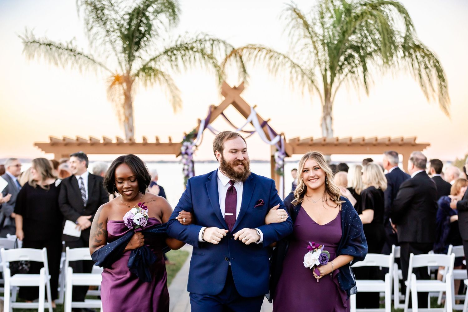 Wedding party members in blue suits and purple dresses walk down outdoor ceremony aisle.