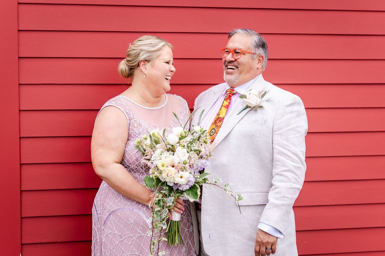 A couple shares a moment in front of a bright red wall, wearing coordinated formal attire.