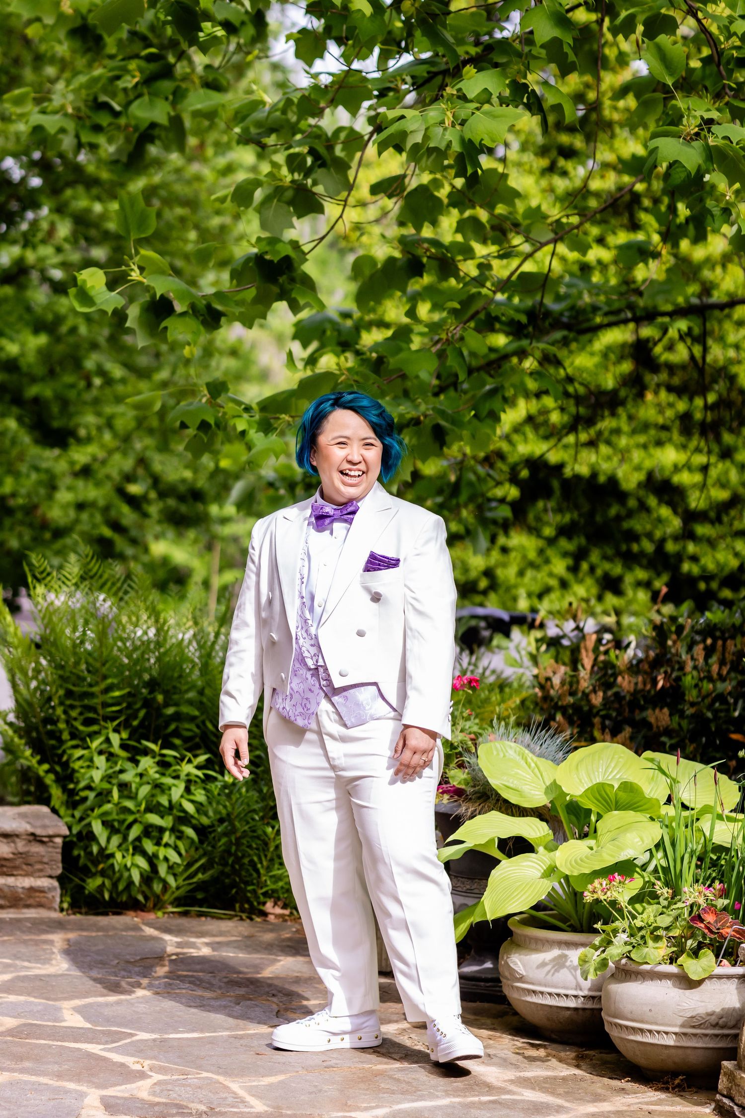 Person in white suit and purple accessories stands on stone patio surrounded by greenery.