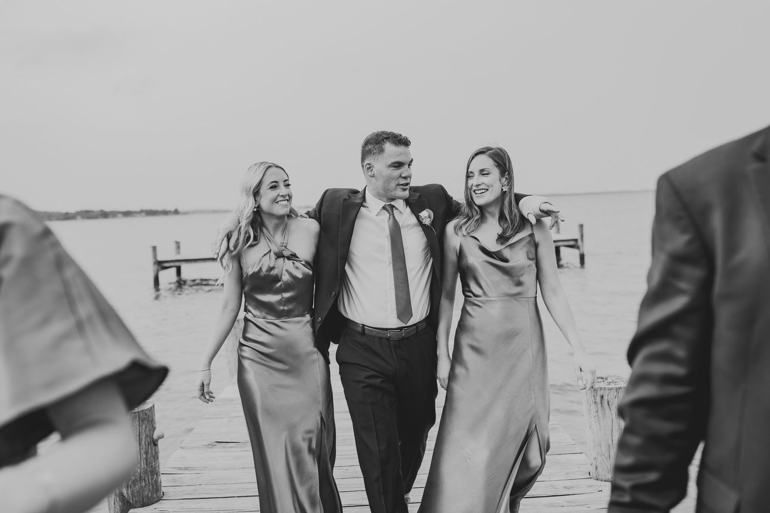 Black and white photo of a wedding party walking together and laughing on a dock near the water.