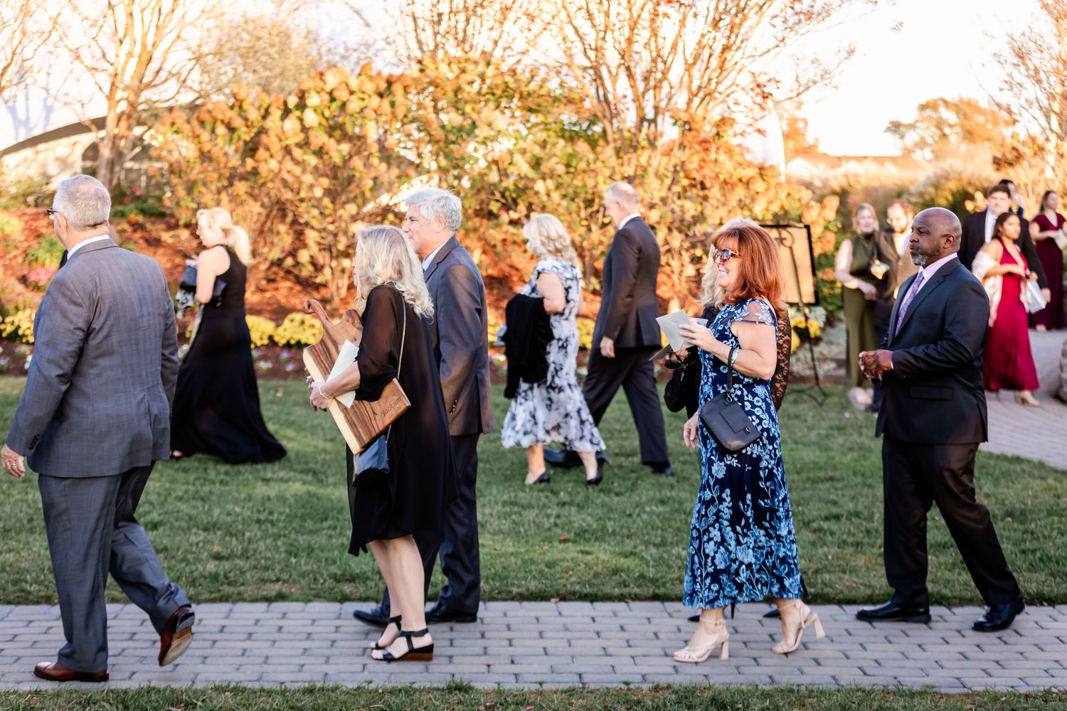 Wedding guests in formal attire walk along a garden path during golden hour at an outdoor reception event.