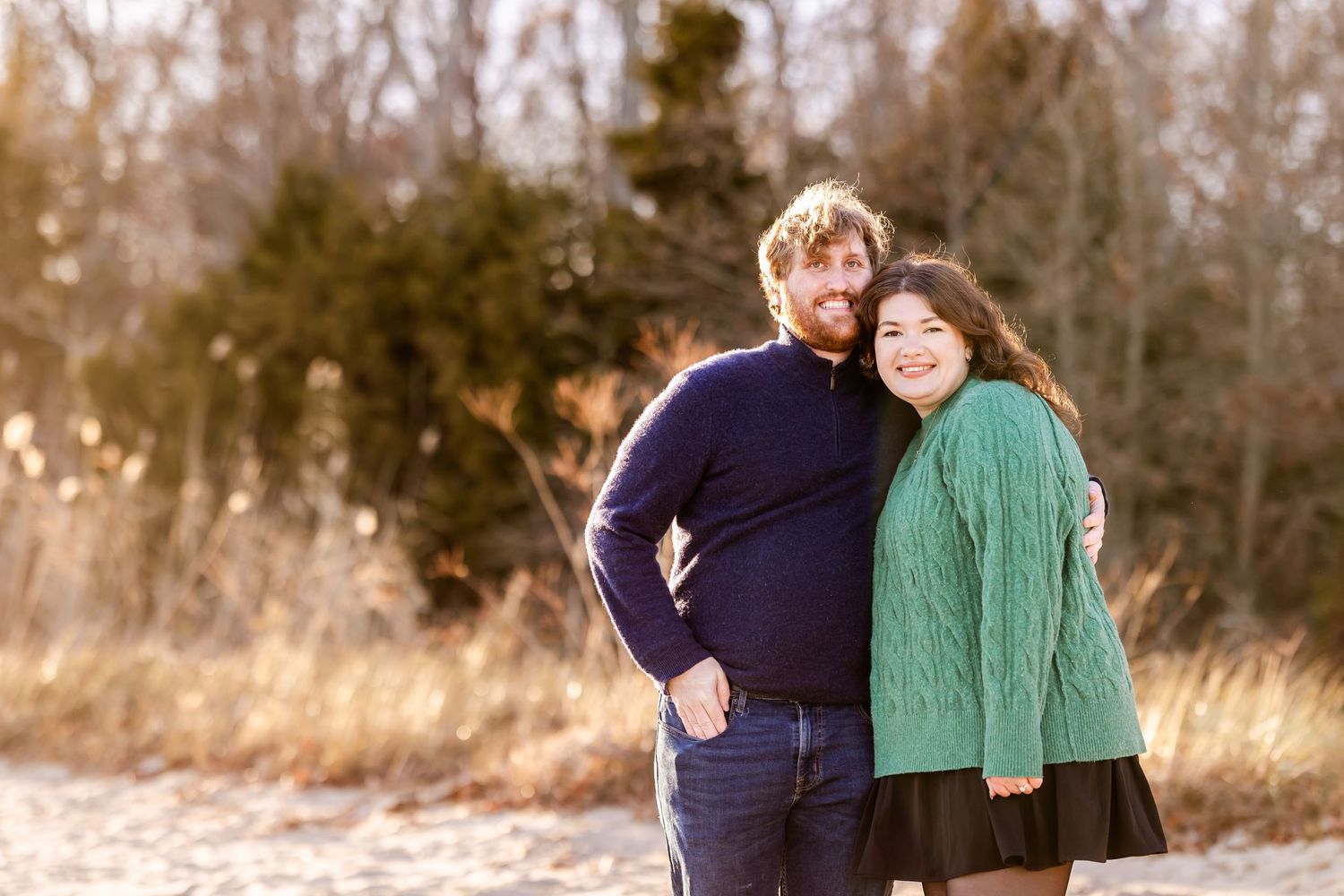Couple in winter clothing embraces while standing in dried grass field during golden hour.