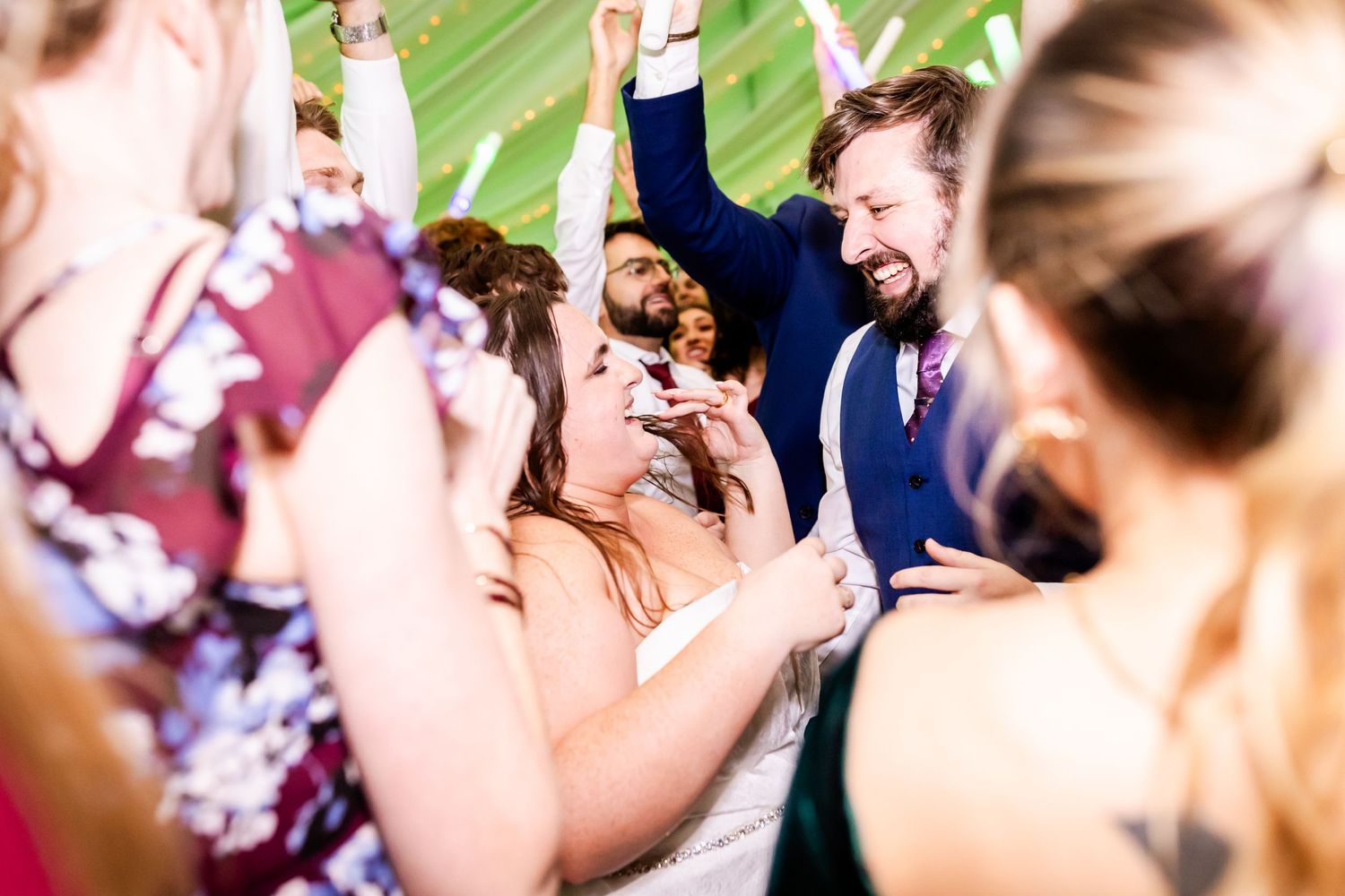 Wedding party celebrates with joyful dancing and laughter under green tent lighting.