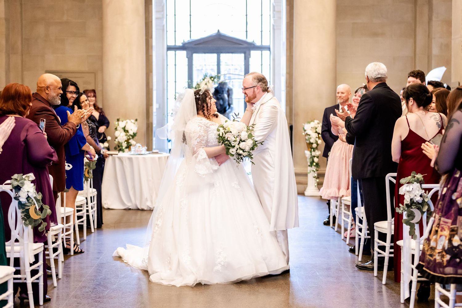 Wedding ceremony with guests watching as couple exchanges vows in a columned hall with floral decor.