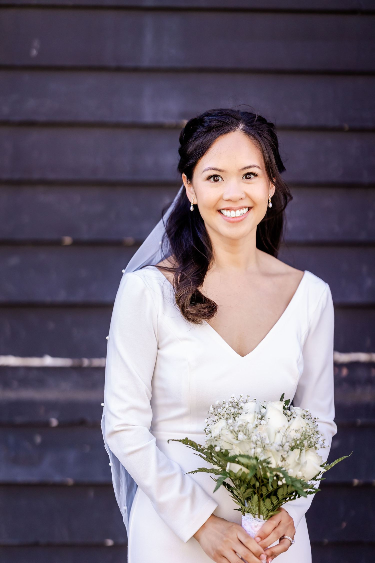 A bride in a long-sleeved white dress holds a delicate bouquet against a dark background.
