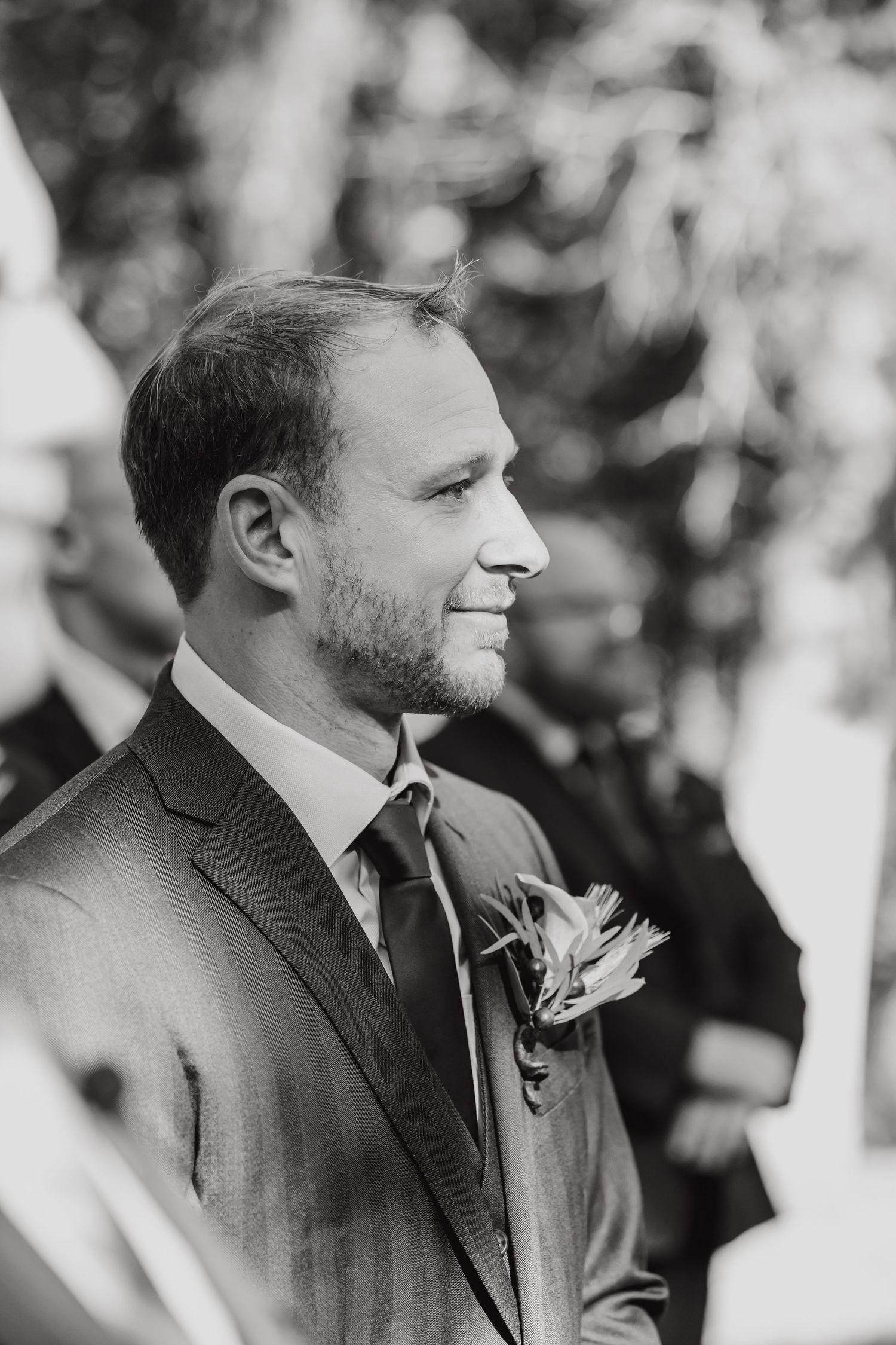 Close-up black and white profile shots of a groom wearing a dark suit with boutonniere at an outdoor wedding.