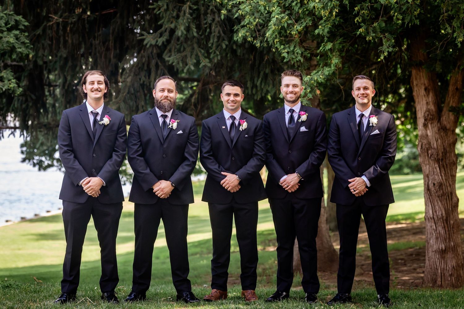 Groomsmen in navy suits pose outdoors with lake and trees in background.