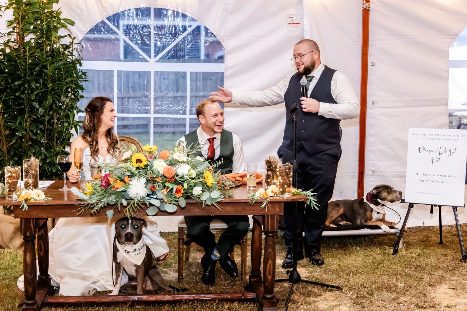 Wedding reception setup features rustic wooden tables and chairs under an elegant white tent with greenery.