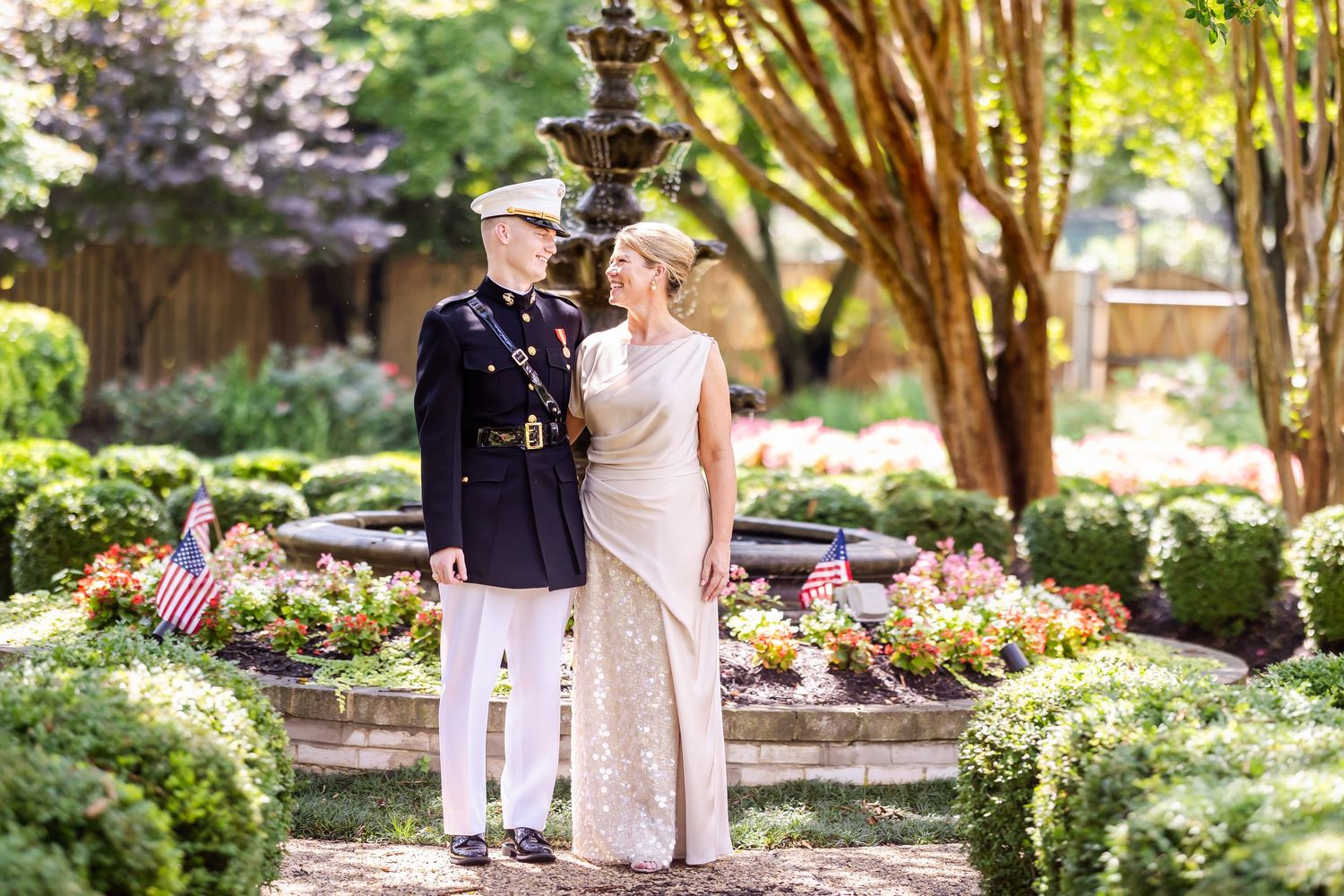 Military couple poses for romantic portrait in front of garden fountain surrounded by flowers.