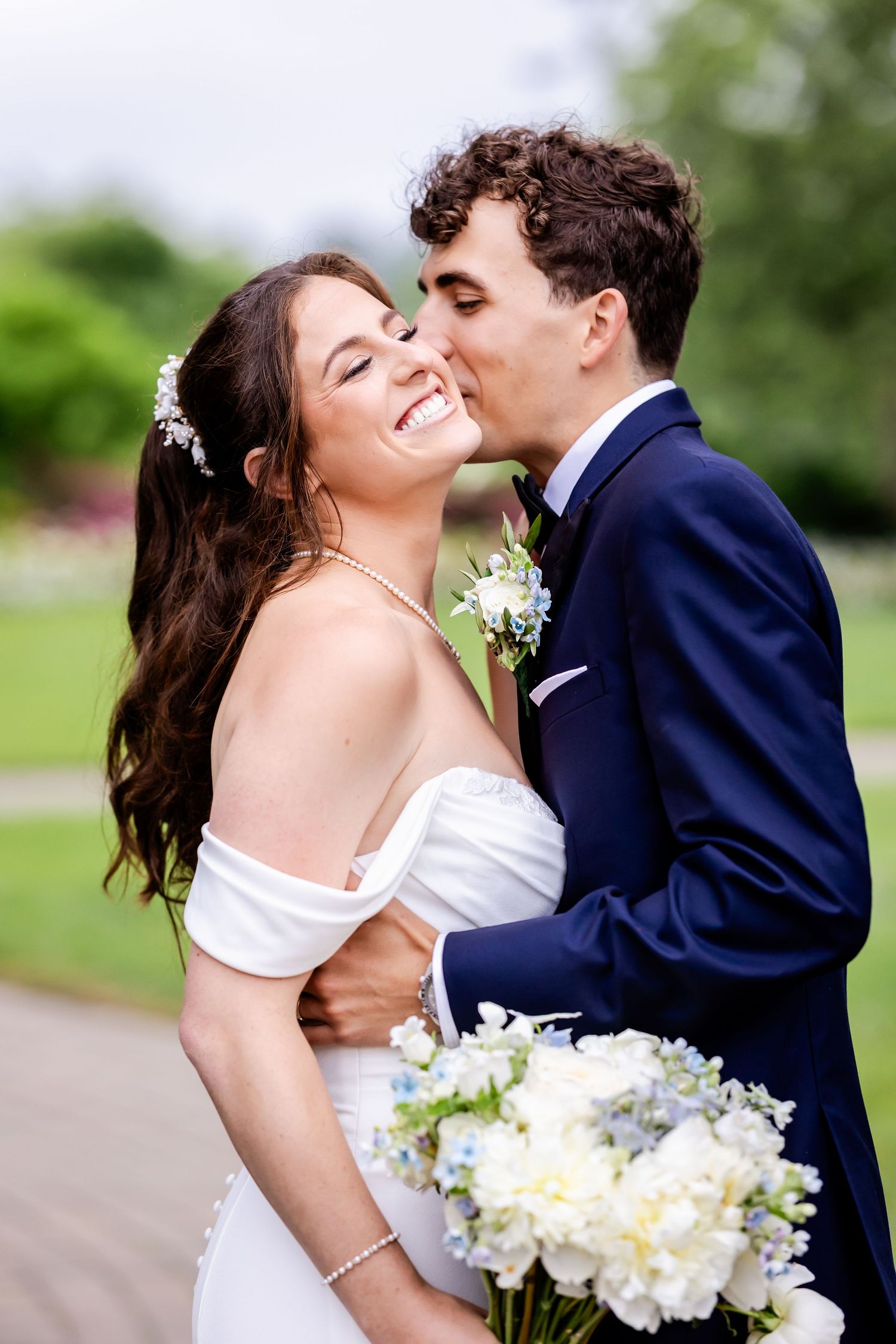 A romantic wedding couple shares a tender kiss while holding a white floral bouquet in an outdoor garden setting.
