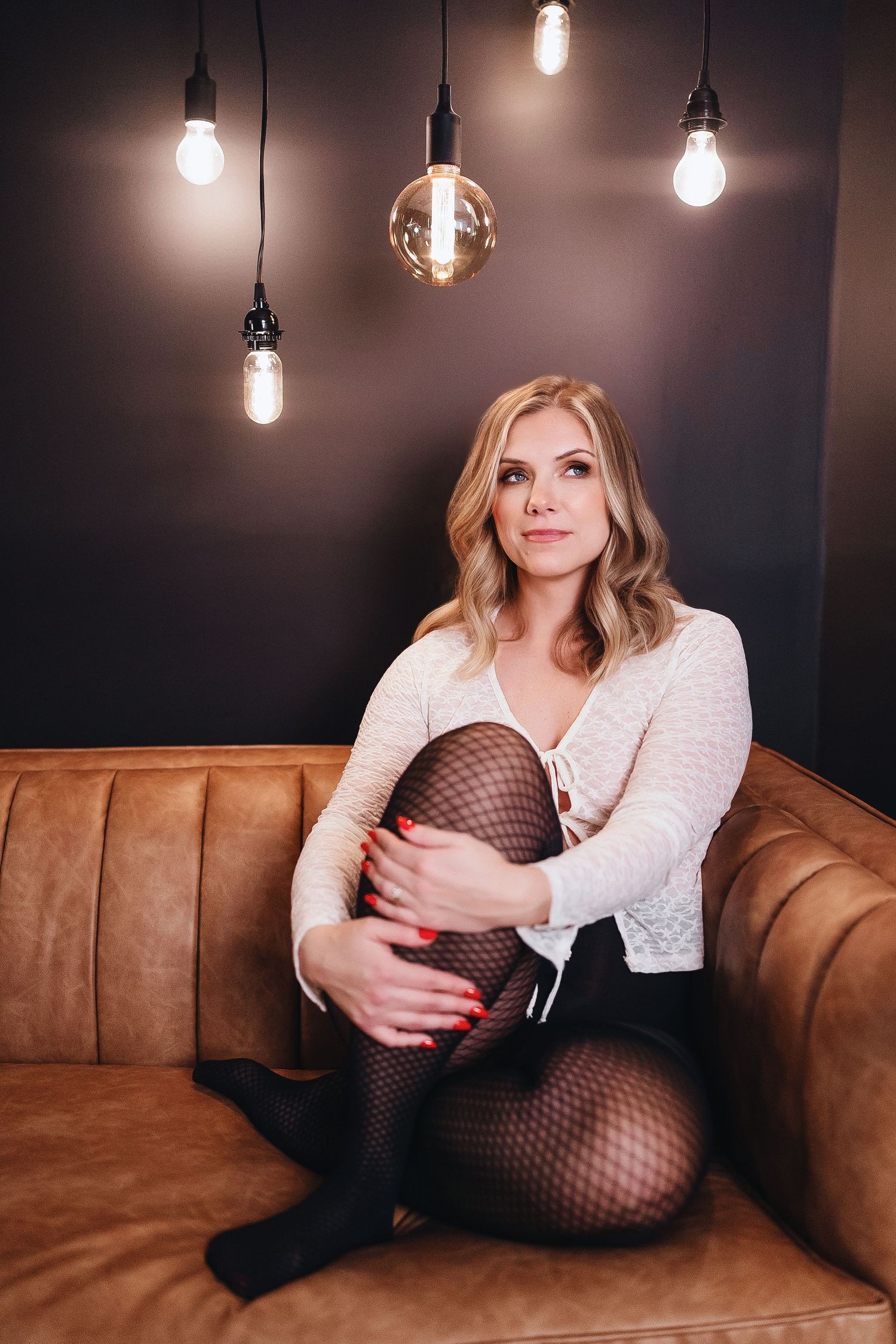 A person in a white cardigan and fishnet stockings poses on a brown leather couch under hanging pendant lights.