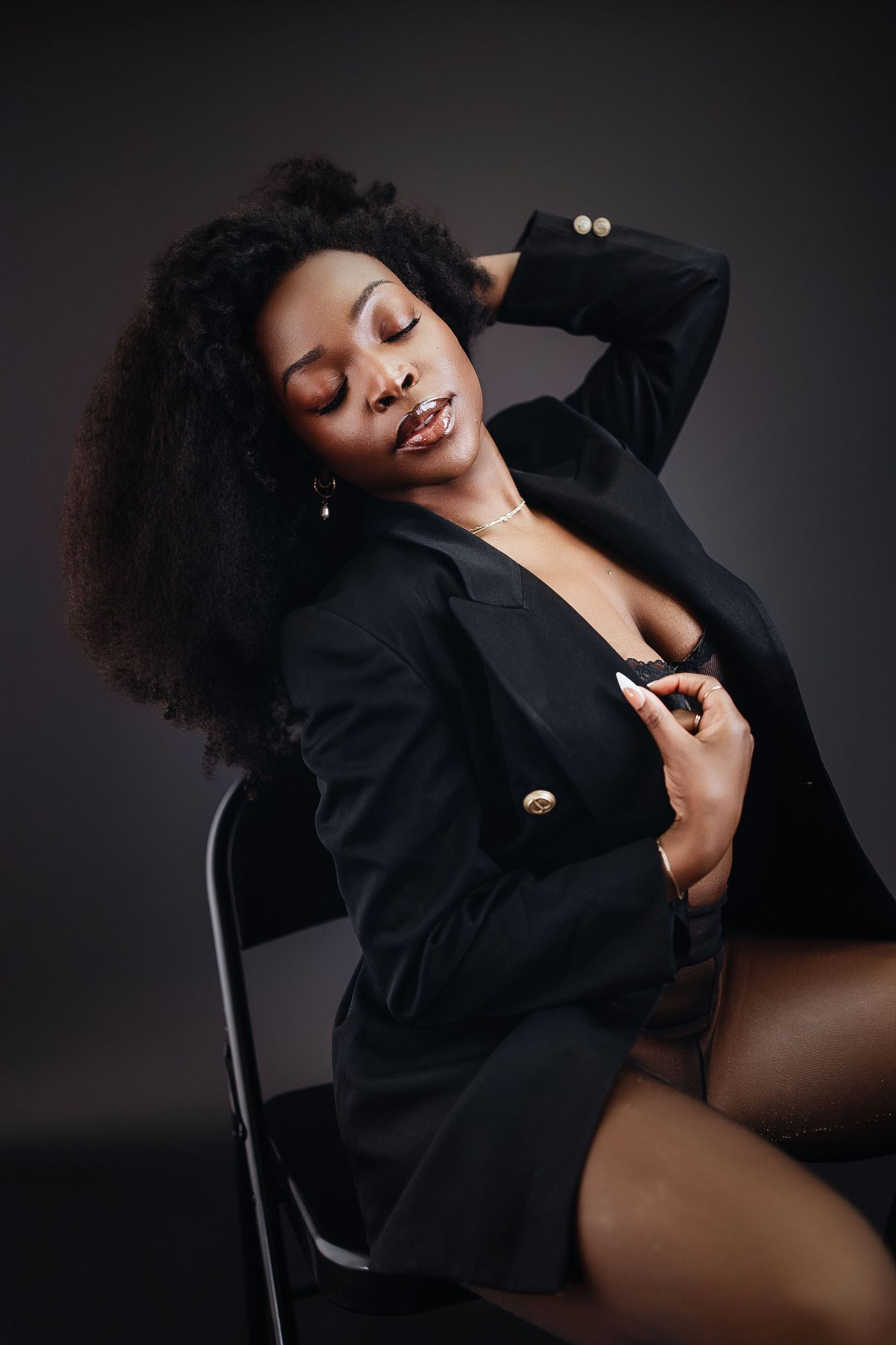 A confident woman in a black blazer poses gracefully on a chair against a dark studio background.