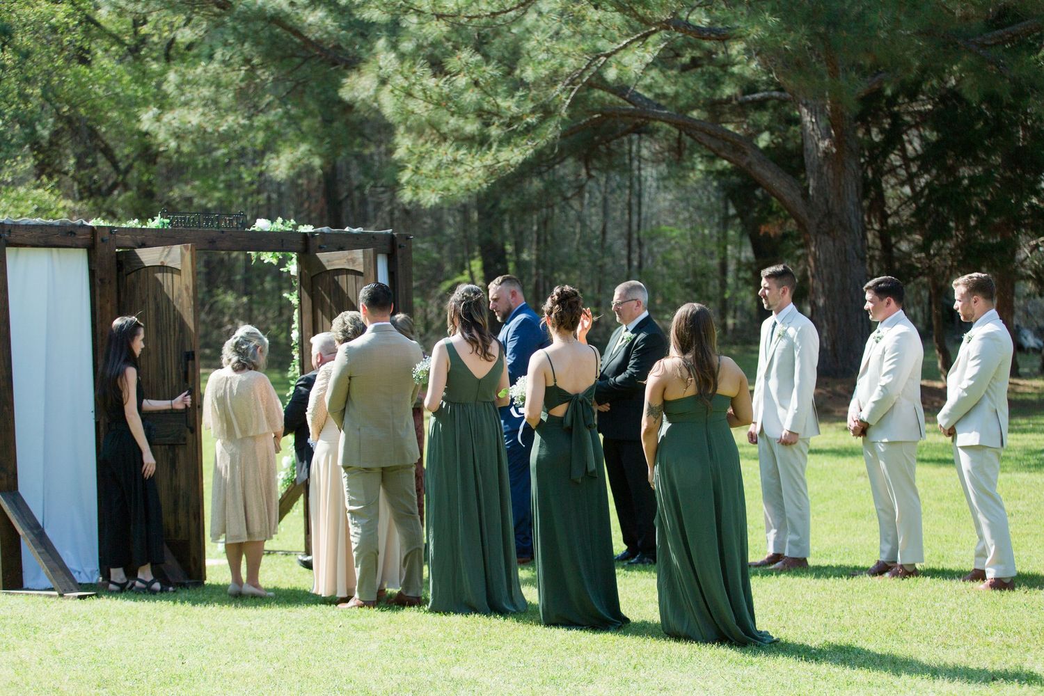A wedding party lines up outdoors on a sunny day with bridesmaids in green dresses and groomsmen in light colored suits.