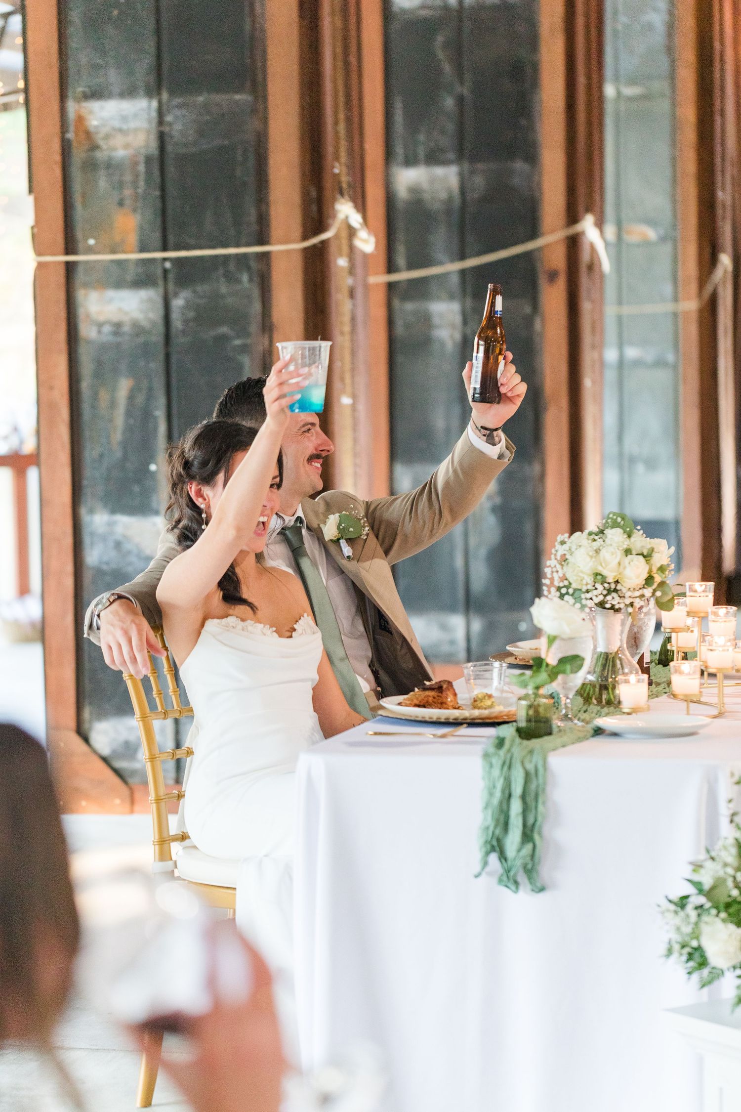 Wedding reception guests celebrating and raising their hands at a long dining table decorated with flowers.